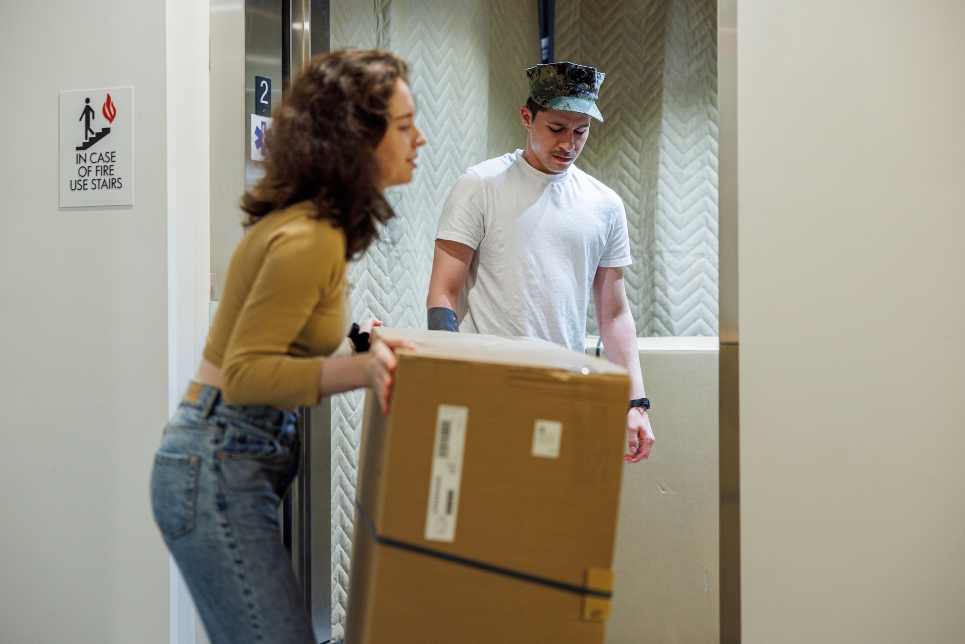 Moving in. Latin man, a navy soldier, and Caucasian woman carrying heavy cardboard boxes into the elevator in the entrance hall.